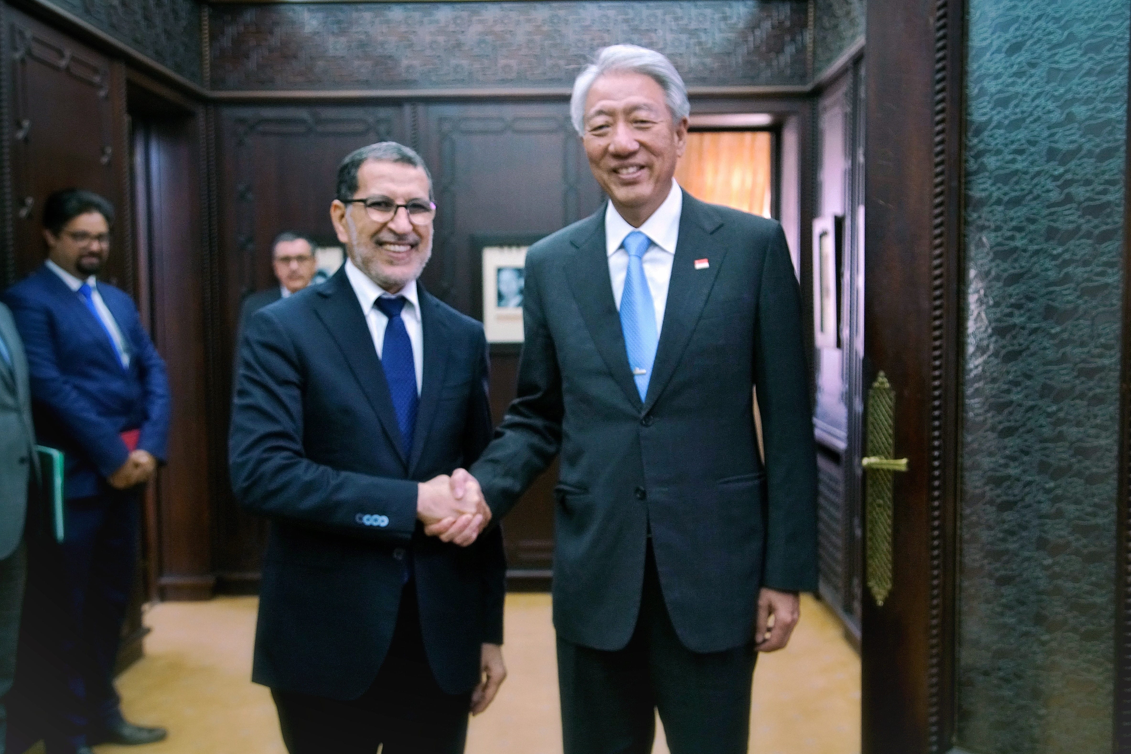 Two men in suits shake hands in a room with wood paneled walls. Man on right wears Indonesian flag pin.